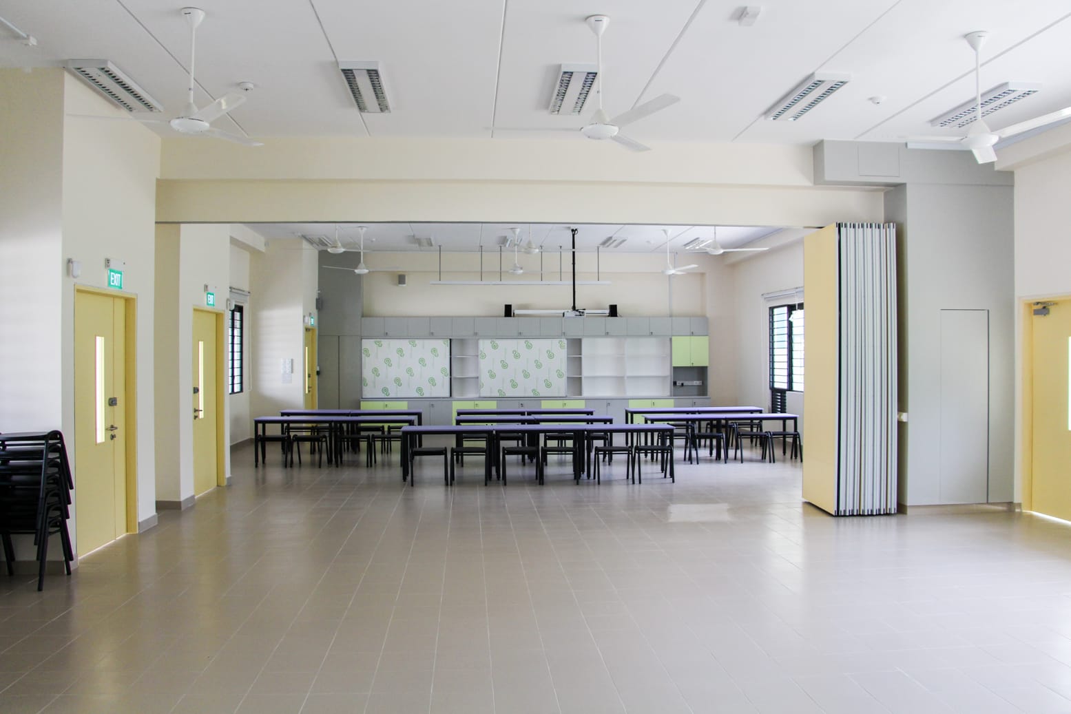 Large empty hall with rows of black tables and chairs, ceiling fans, yellow doors, and storage cabinets at the back.